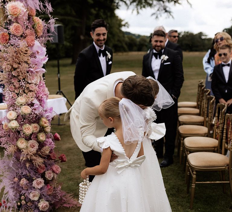 Groom in white tux saying hello to his little girls in white flower girl dresses with bows 