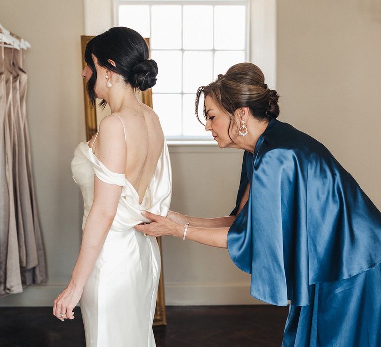 Mother of the bride in a blue satin dress helps the bride into her wedding dress 