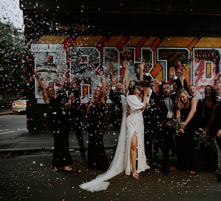 The wedding party celebrates the wedding with confetti as bride and groom share a kiss 