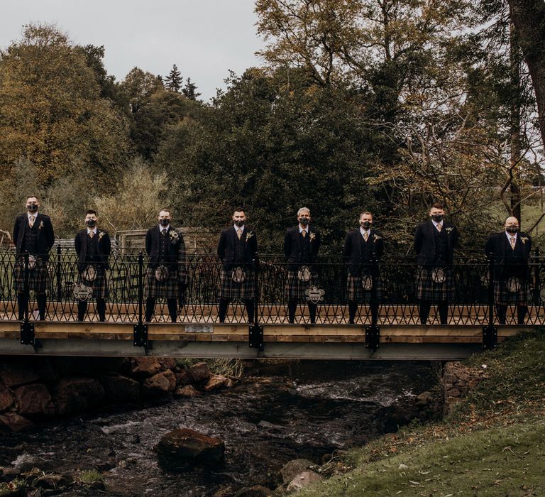 Groomsmen in kilts pose on bridge wearing facemasks at highland wedding in Glencoe 