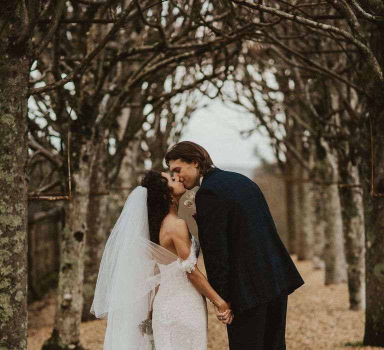 Groom in a navy suit leaning down and kissing his bride in a fitted lace wedding dress by Pronovias 