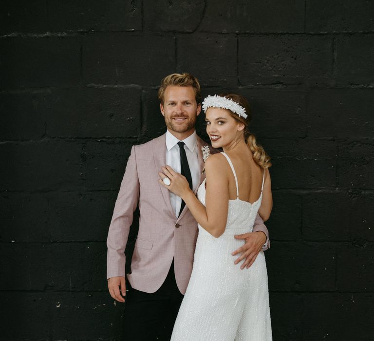 Groom in pink jacket embracing his bride in a wide leg sequin jumpsuit and headband 