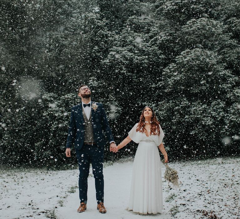 Bride in white dress holding dried flower bouquet holds hands with groom in navy suit and tartan waistcoat during snowfall at Cannon Hall