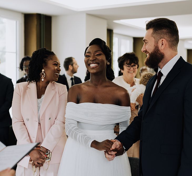 Black bride wears off the shoulder wedding gown holding hands and smiling with groom