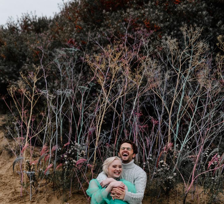 Groom in a grey jumper sitting in the sand embracing his bride in a sheer pastel wedding dress for intimate beach elopement 