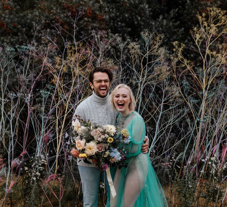 White blonde bride with bob length hair laughing on the beach in a sheer pastel wedding dress with her bearded groom in a grey sweater