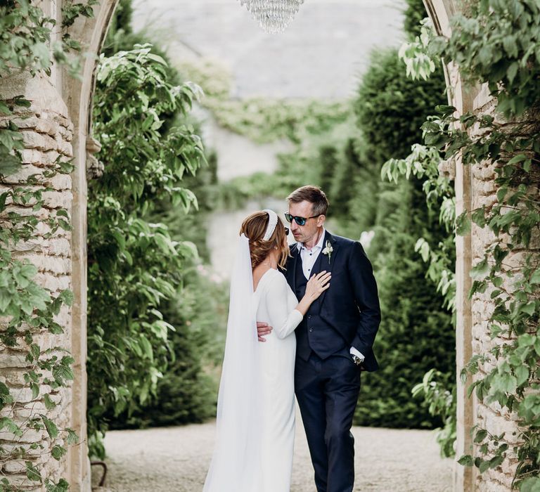 Bride in Elbeth Gillis gown kisses groom in navy Cad & the Dandy suit and sunglasses under chandelier hanging in archway outside at Euridge Manor