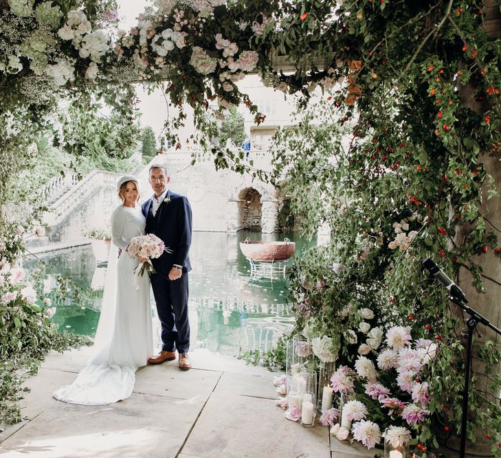 Bride in Elbeth Gillis gown holding blush pink rose wedding bouquet stands outside with groom in navy Cad & the Dandy suit under floral canopy at Euridge Manor wedding