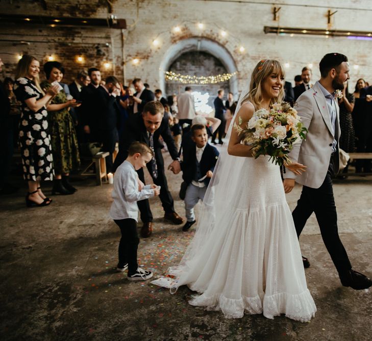 Bride and groom cross hall as guests applaud. Bride wearing white wedding dress, veil and carrying bouquet. Groom wearing black trousers and grey jacket. 