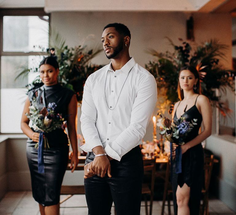 Groom in leather trousers, white open buttoned shirt and silver necklace stood with bridesmaid in leather dresses 