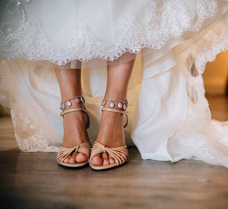 Bride wearing beige strappy bridal shoes with pearl ankle bracelets and lace bridal gown