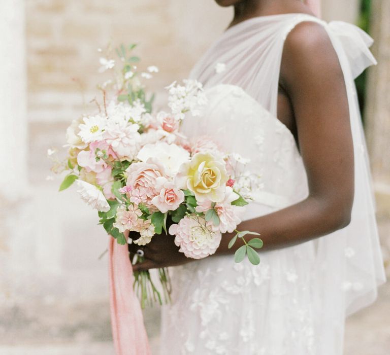 The bride holds a bouquet of pink roses, dahlias and carnations wearing a one shoulder Anna Campbell wedding dress with pink ribbon in her hair