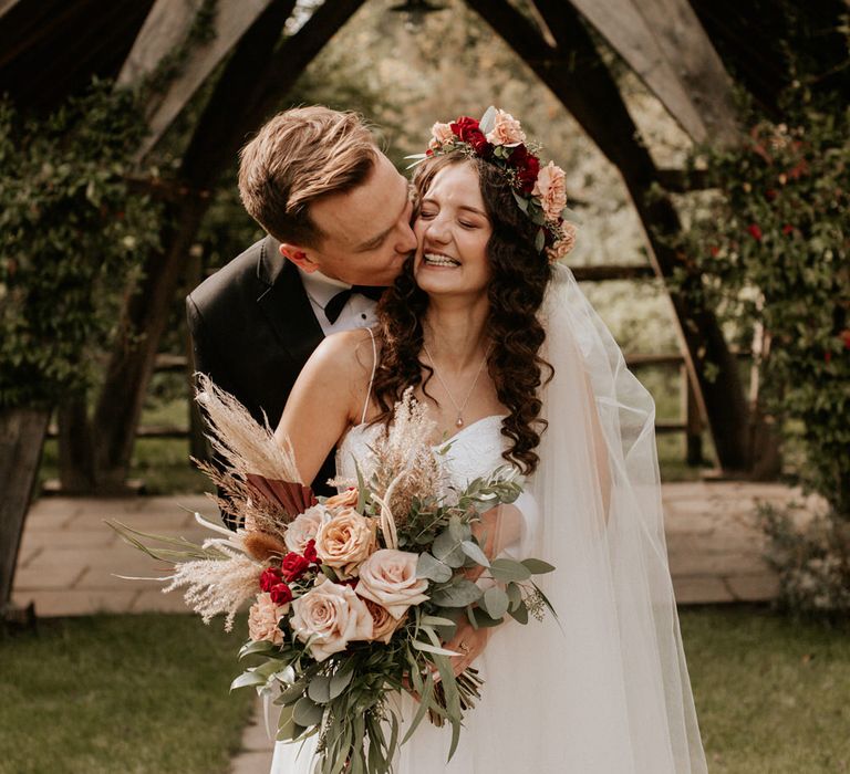 Groom kissing his boho bride with long wavy hair and red and green flower crown