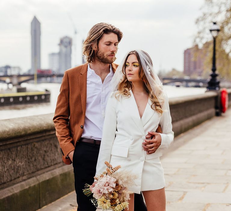 Stylish bride in orange suede platforms with floral socks and blazer dress standing with her groom by the Thames in Chelsea