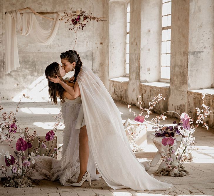 Bride with braided wedding hair wearing a bridal cape and white stud heels leaning her bride down for a kiss amongst purple anthurium wedding flower displays