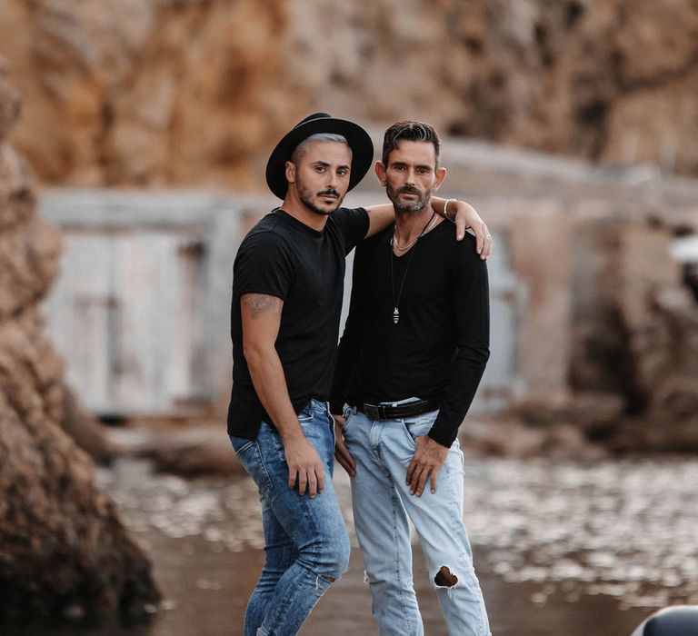 Grooms stand together wearing black T-shirts and pale blue jeans on the beach