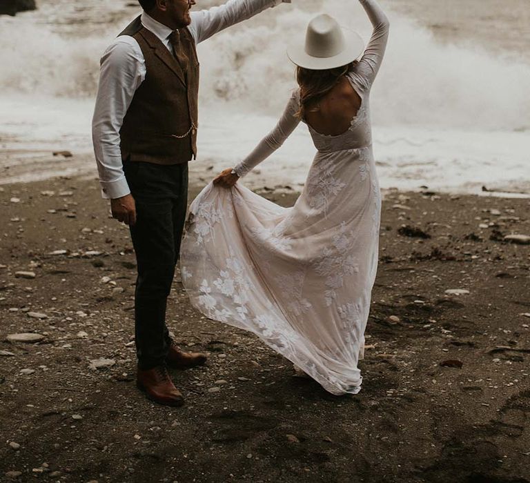 Groom in a check waistcoat twilling his bride on the beach in a fitted wedding dress and hat 