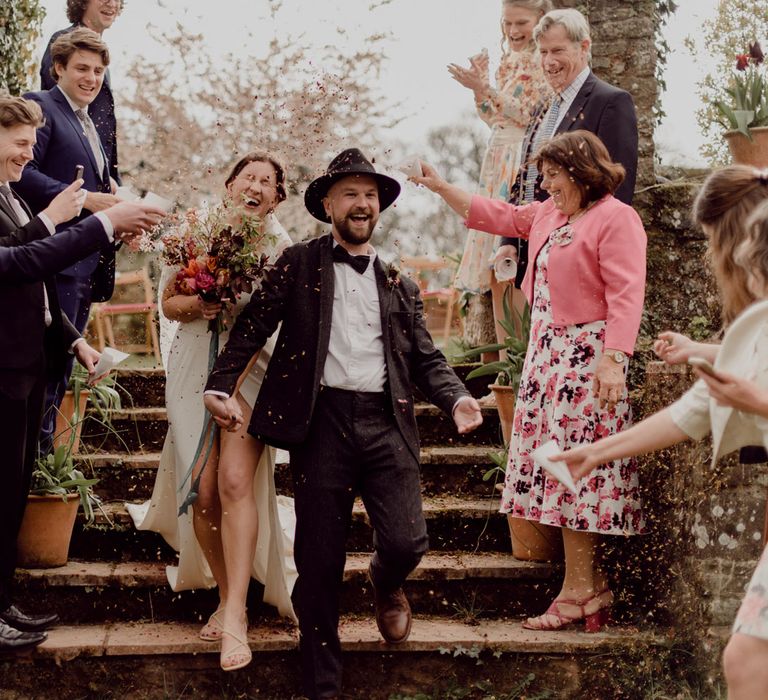 Laughing groom in grey tweed suit and black fedora holds hands with laughing bride in Charlie Brear wedding dress and applique veil walking down garden steps whilst guests throw confetti at garden party wedding in Devon