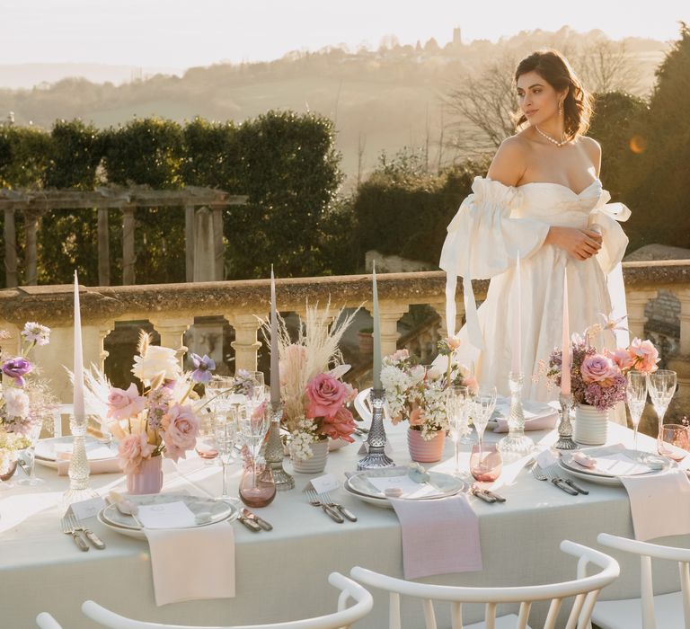 Bride stands behind luxury wedding tablescape whilst the sun shines and pastel floral decor lines the table 