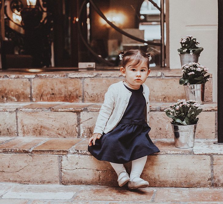 Little guest in a navy dress and silver cardigan sitting on some steps with silver bucket plants 