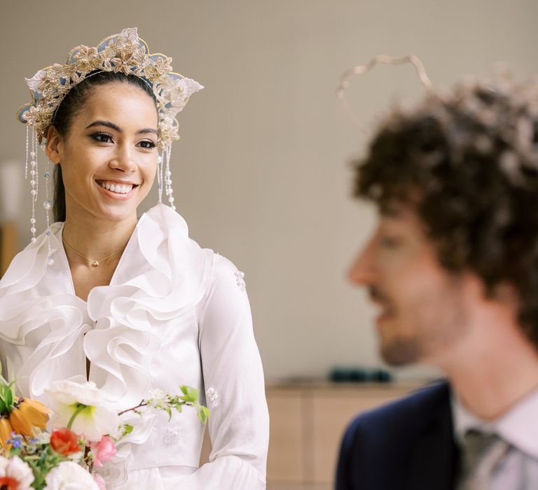 Bride smiles sweetly at groom whilst wearing golden embroidered crown and holding pastel floral bouquet