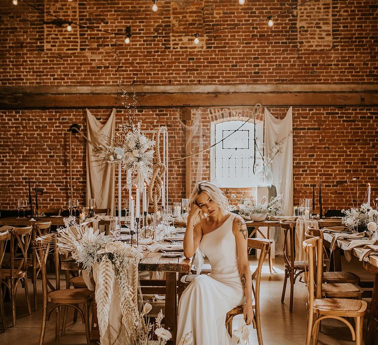 Stylish bride in a one shoulder wedding dress with puddle train sitting in a bohemian luxe wedding reception at Godwick Great Hall and Barn