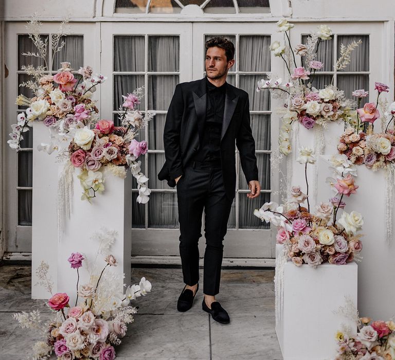 Stylish groom in a black shirt, loafers and tuxedo standing next to a display of romantic pink, lilac white and peach peeled back roses 