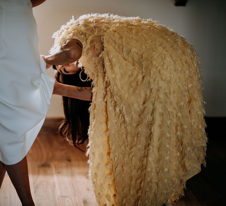 Bride in white Rebecca Vallance dress stands whilst guest in yellow feather detail dress adjusts the hem before Harrogate wedding