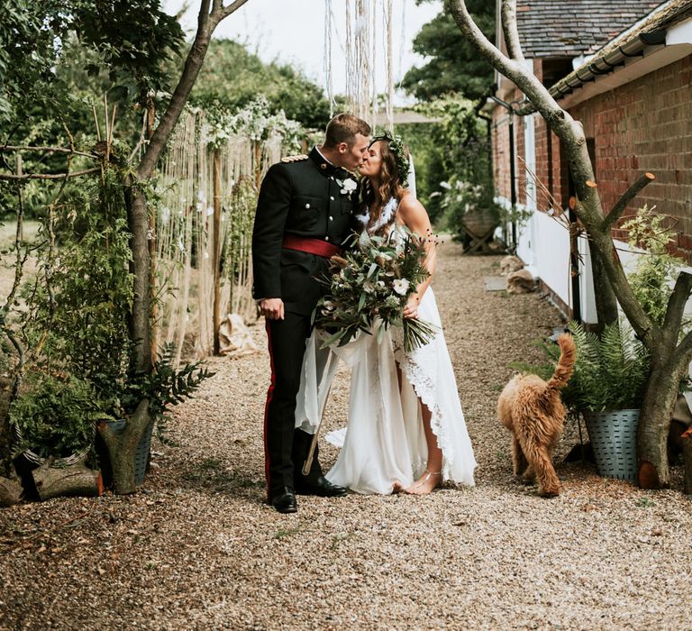 Bride in a Hollie Grace Loves Lace boho wedding dress kissing her groom in military uniform under a giant dream catcher 