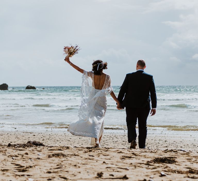 Bride celebrates and holds dried floral bouquet in the air as she walks along the beach with her groom