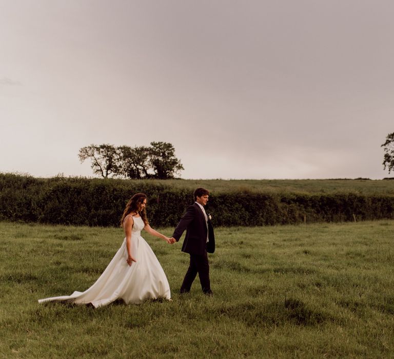 Bride in white Elbeth Gillis wedding dress walks across field holding hands with groom in dark brown suit at home farm wedding