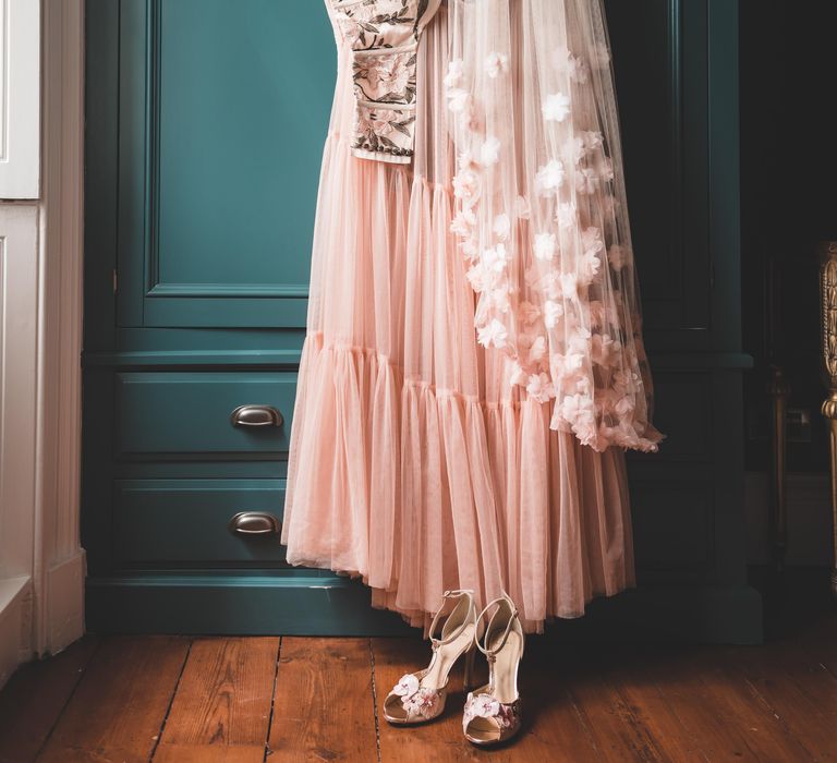 Blush pink appliqué veil hanging up with an embroidered bodice, tulle skirt and T-bar wedding shoes 