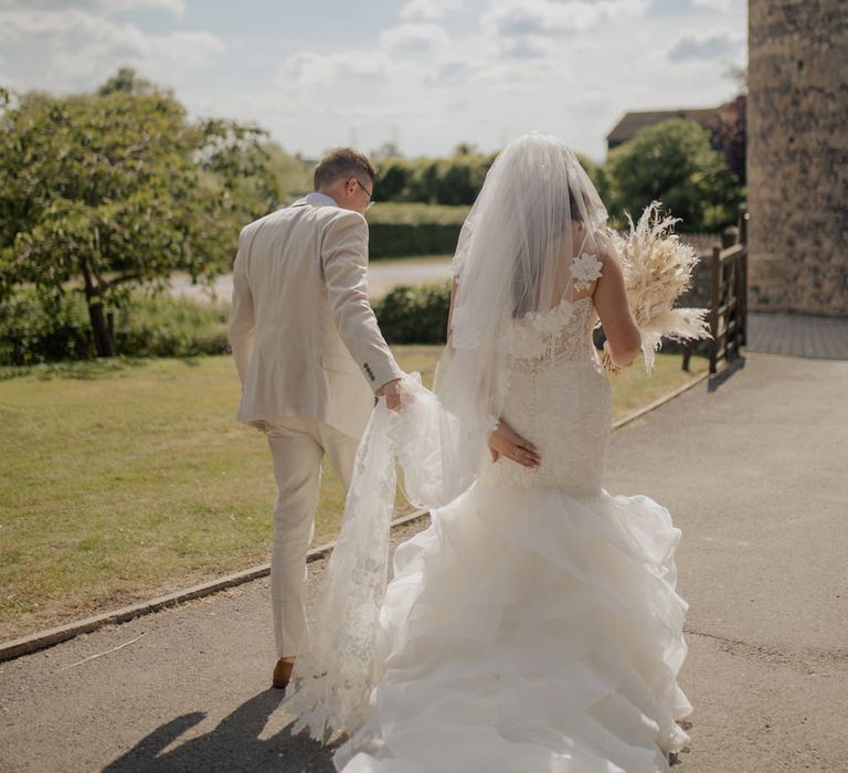 Groom in a beige suit holding his brides appliqué wedding veil as she walks along in her fishtail wedding dress