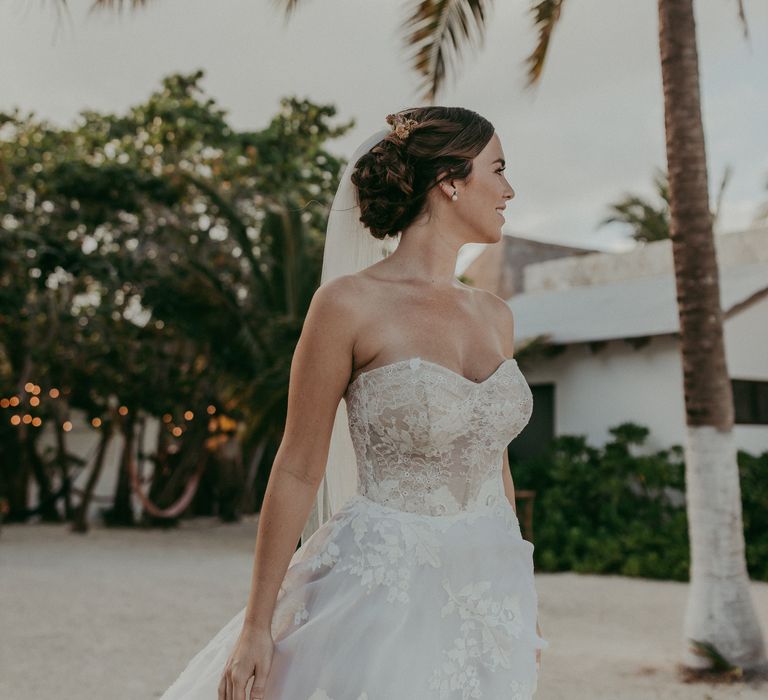 Bride stands beside palm tree as she walks along the beach during her wedding day