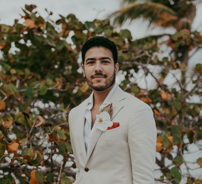 Groom looks toward camera as he wears sand coloured suit, red pocket square and orchid buttonhole 