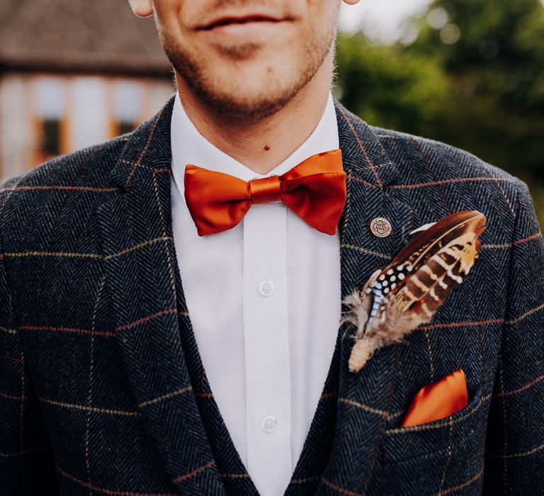 Groom wears orange bow-tie and suit features orange threading throughout, along with orange pocket square and feather buttonhole