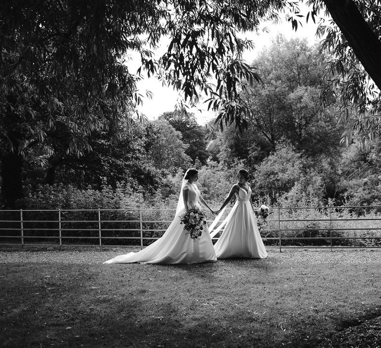 Bride in white cami wedding dress and shoulder veils walks through the grounds of The West Mill Derby holding hands with bride in lace top capped sleeve wedding dress and veil 