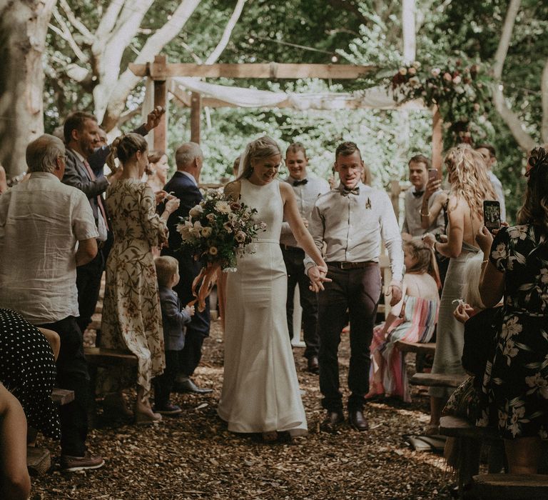 Bride & groom walk through Lila's Wood after wedding ceremony outdoors under rustic wooden pergola complete with floral decor