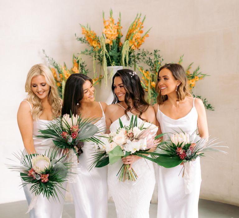 Bridal party in white satin bridesmaid dresses holding tropical flower wedding bouquets 