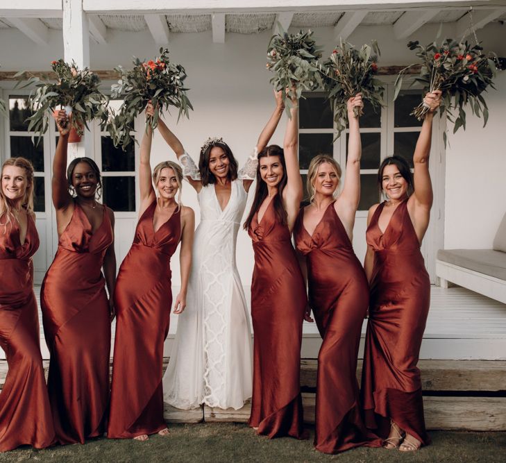 Destination wedding with bride in lace Grace Loves Lace wedding dress and bridesmaids in brown satin halterneck dresses holding their bouquets in the air  