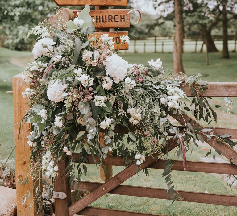 White, pink and green florals and foliage draped over wooden gate next to wooden post sign at Tythe Barn wedding with barn wedding flowers