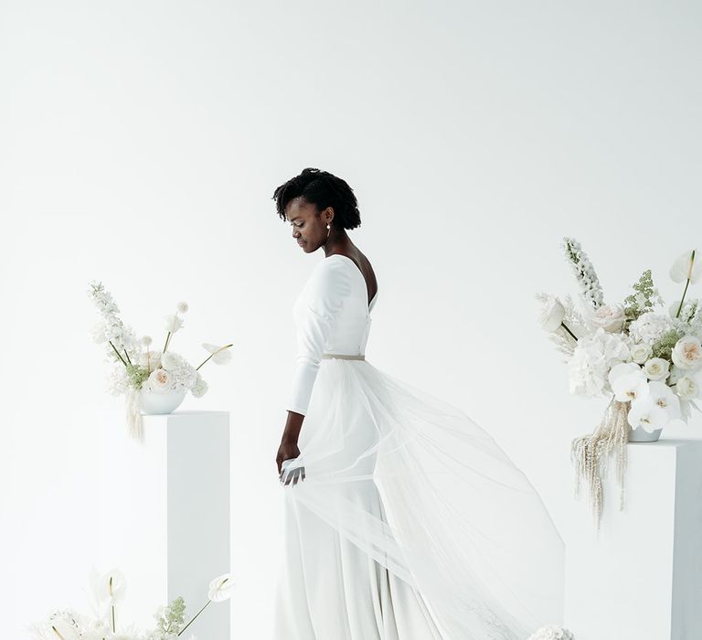 Black bride in a fitted wedding dress with tulle detachable skirt standing in an industrial wedding venue decorated with all white wedding flowers 