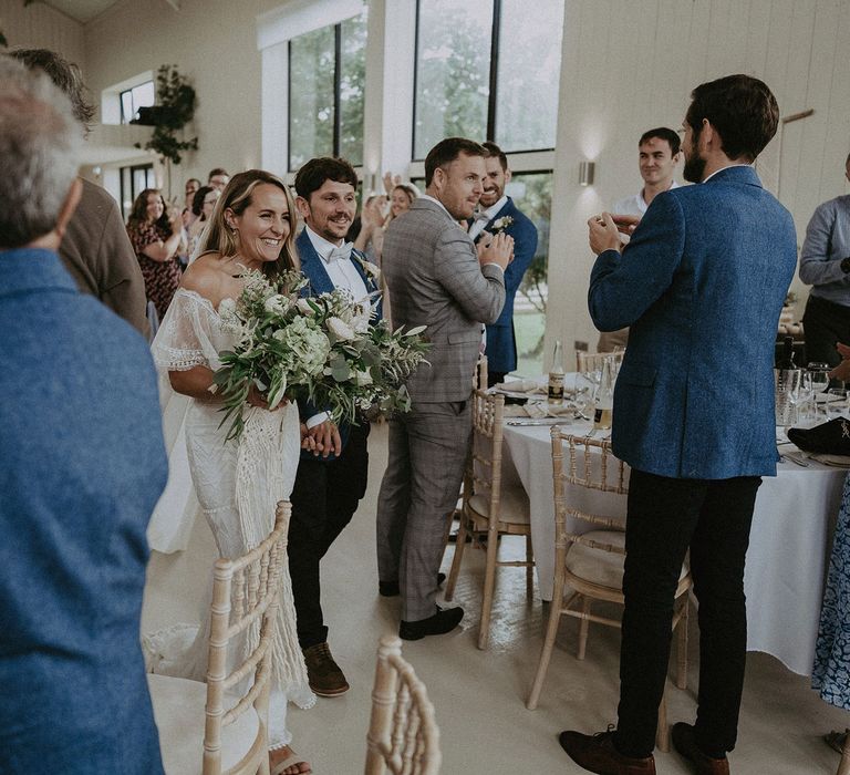 Bride in Grace Loves Lace bardot wedding dress with white and green bridal bouquet walks with groom in blue suit jacket through wedding breakfast at Isle fo Wight wedding with macrame wedding decor