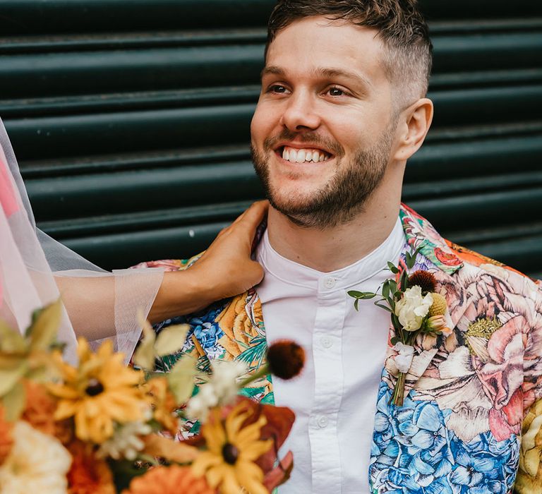 Stylish groom in a white grandad shirt and brightly coloured patterned blazer smiling 