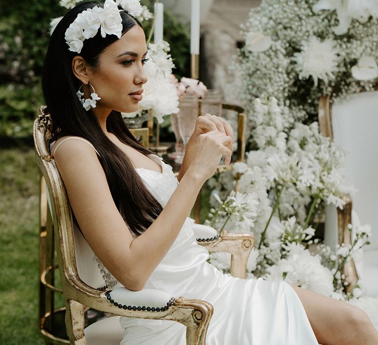 Bride with long brown hair in a white flower headdress wearing a satin and lace wedding dress with front split sitting on a chair 