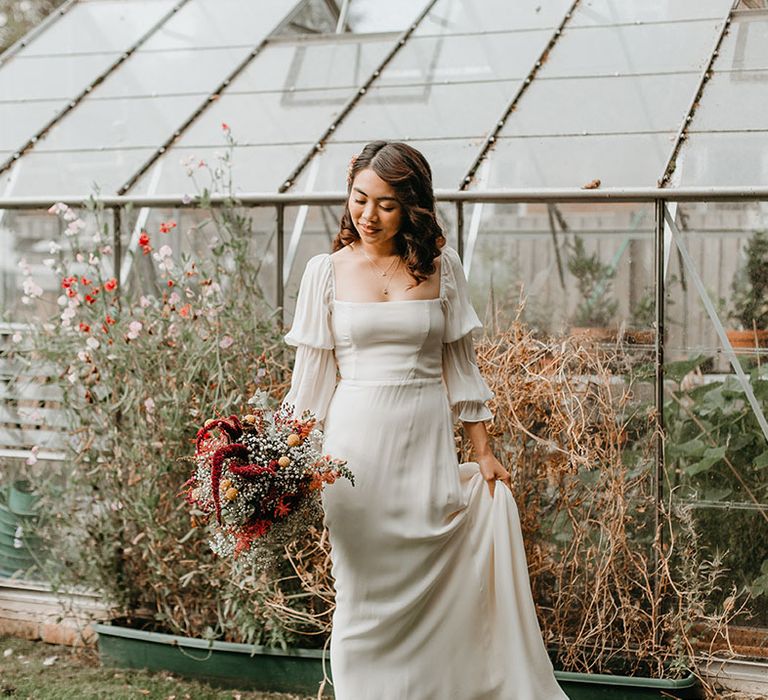 Bride holds her bridal gown complete with long sleeve sheer sleeves and sweetheart neckline whilst holding bridal bouquet