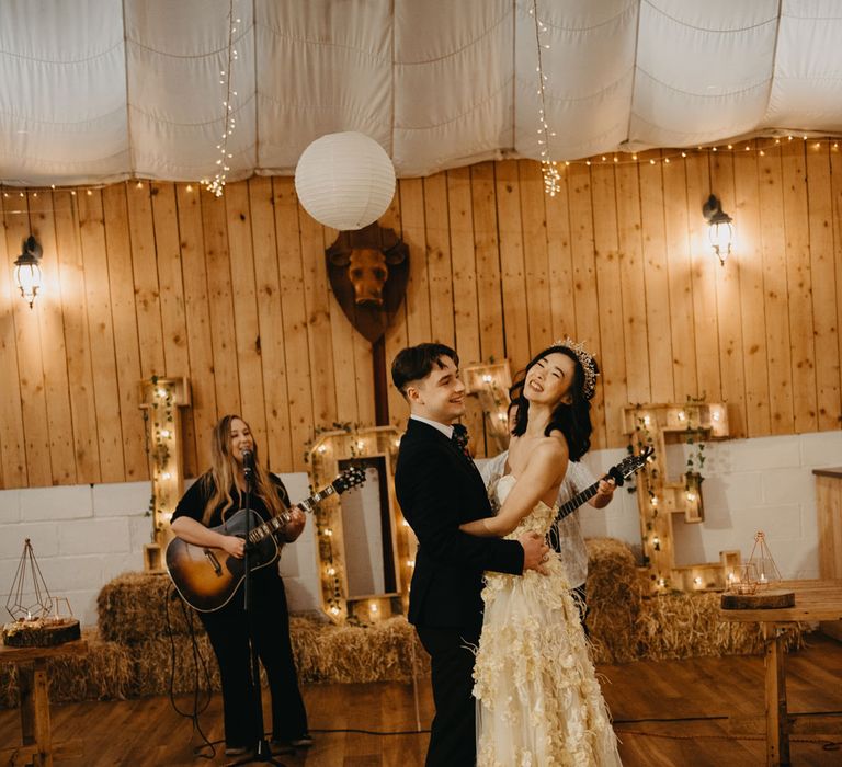 Bride in applique wedding dress and headpiece dances with groom in dark suit whilst guitarists play for rustic wedding reception