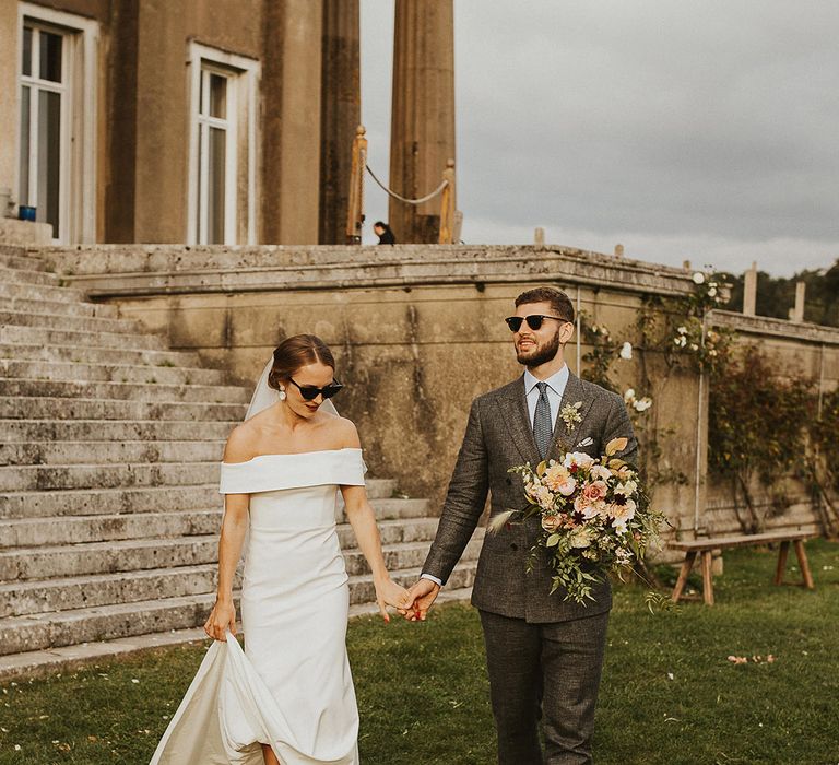 Bride & groom walk hand in hand across lawn on wedding day