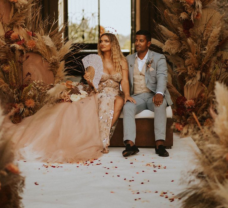 Bride & groom sit together on their wedding day in front of large floral arrangement 