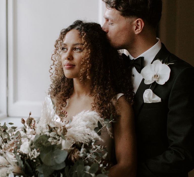 Groom kisses brides head as they stand beside bright window